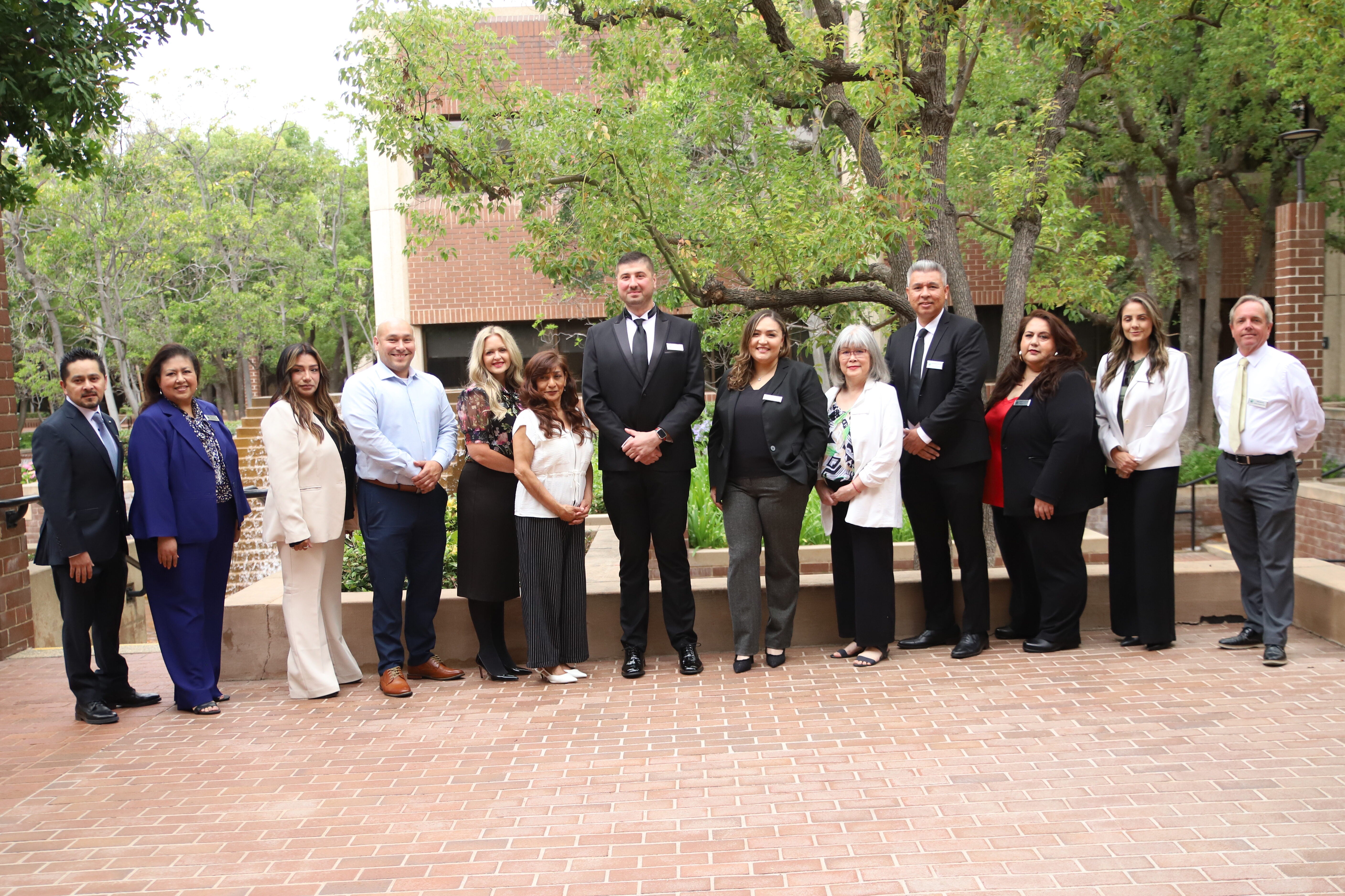 Thirteen people in formal business attire stand outdoors on a brick walkway, posing for a group photo with trees and buildings in the background.