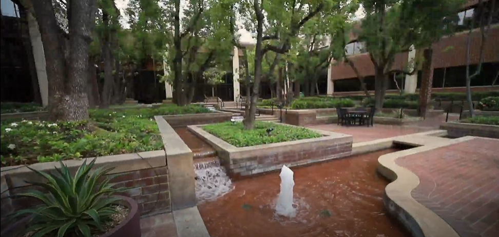 A courtyard with trees, brick planters, a small fountain, and seating areas surrounded by buildings.