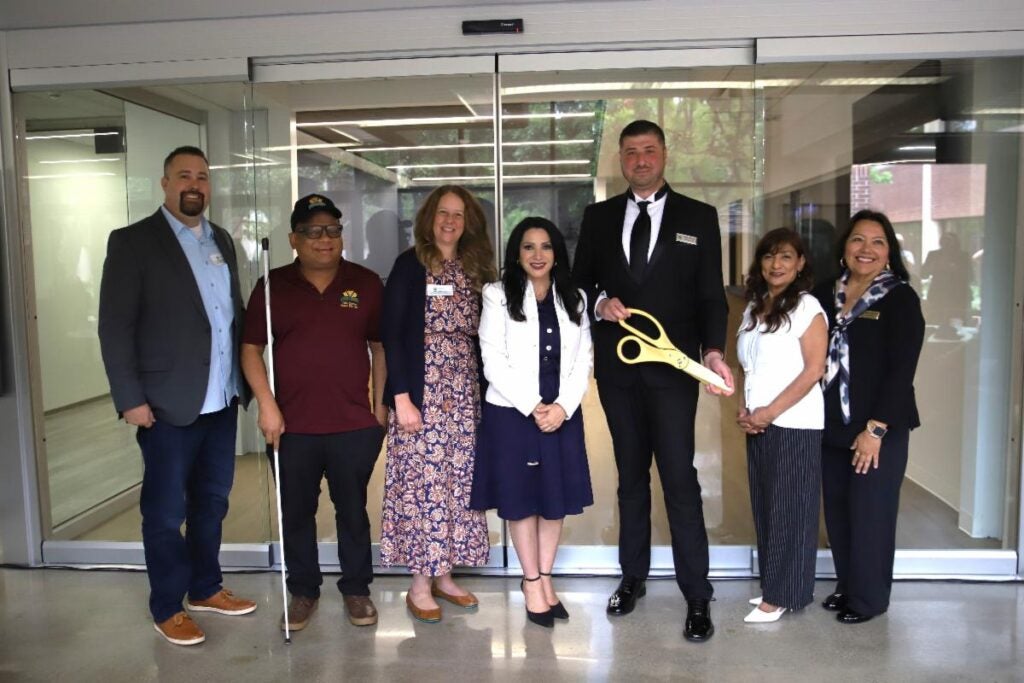Seven people stand indoors in front of glass doors; one person holds oversized ceremonial scissors, suggesting a ribbon-cutting event.