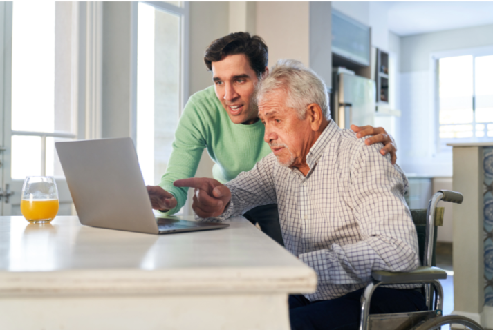 A younger man helps an older man in a wheelchair use a laptop at a kitchen table. An orange juice glass sits nearby.