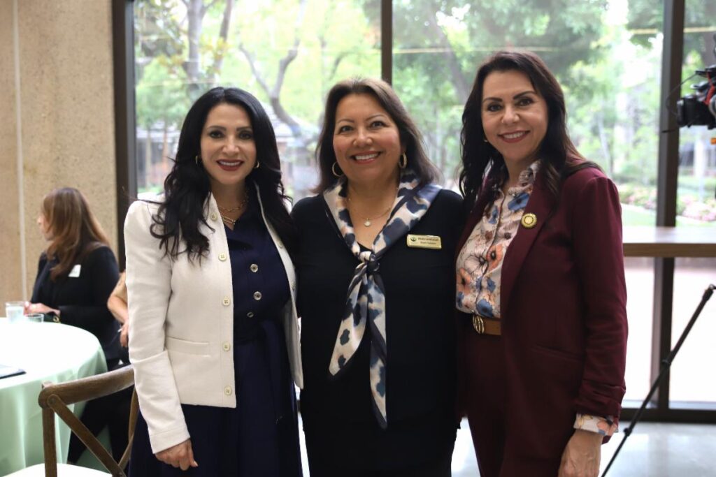 Three women in business attire stand together and smile for a photo at an indoor event with large windows and greenery outside.