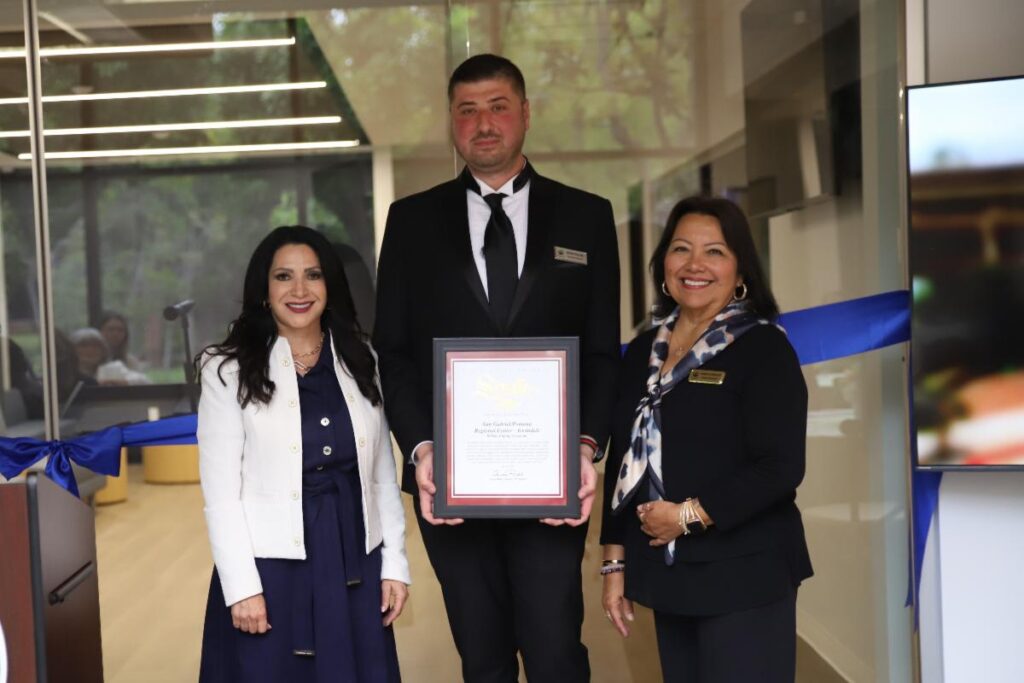 Three people stand indoors, with the man in the center holding a framed certificate. A blue ribbon is stretched behind them, suggesting a ribbon-cutting or award ceremony event.