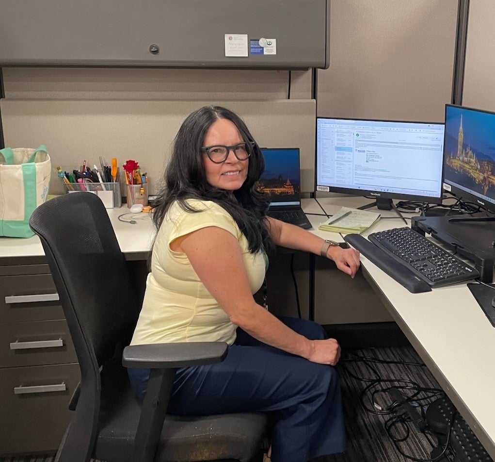 Woman with long dark hair and glasses sits at a desk in an office cubicle, working at a computer with two monitors. Office supplies and a tote bag are visible in the workspace.